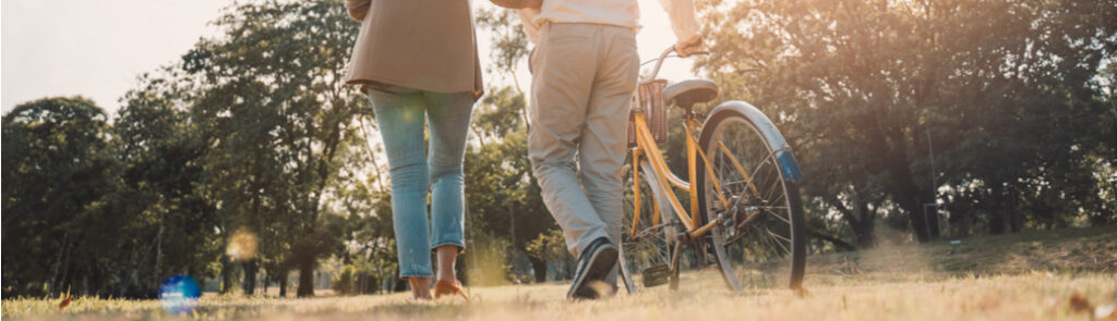 An older couple walking through a park as one pushes a bike.