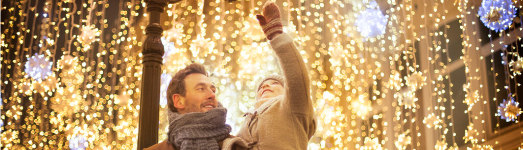 A father holds his young daughter in his arms as she reaches out towards hanging Christmas lights